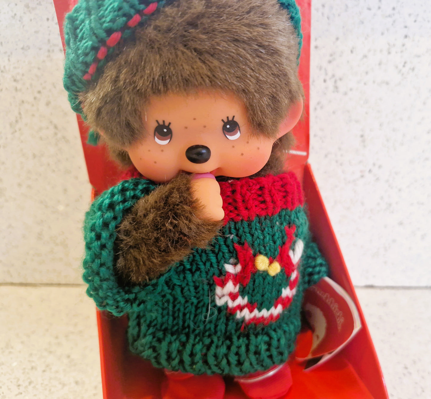 Toy figure wearing a green sweater with a red collar, sitting on a red surface.