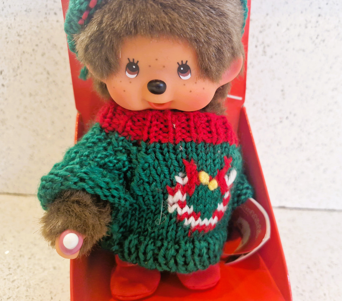 Monchhichi Thumb Sucking Plush, wearing a christmas sweater with a wreath design on a red background