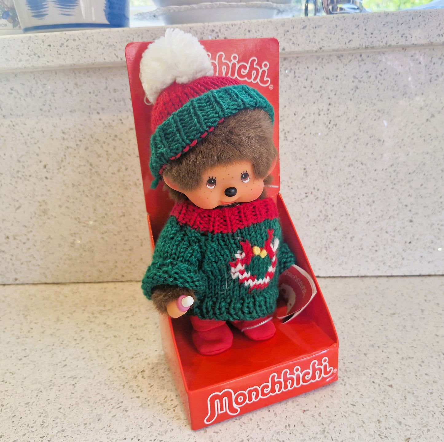 Monchhichi toy in a green outfit with a red sweater on a red packaging box.
