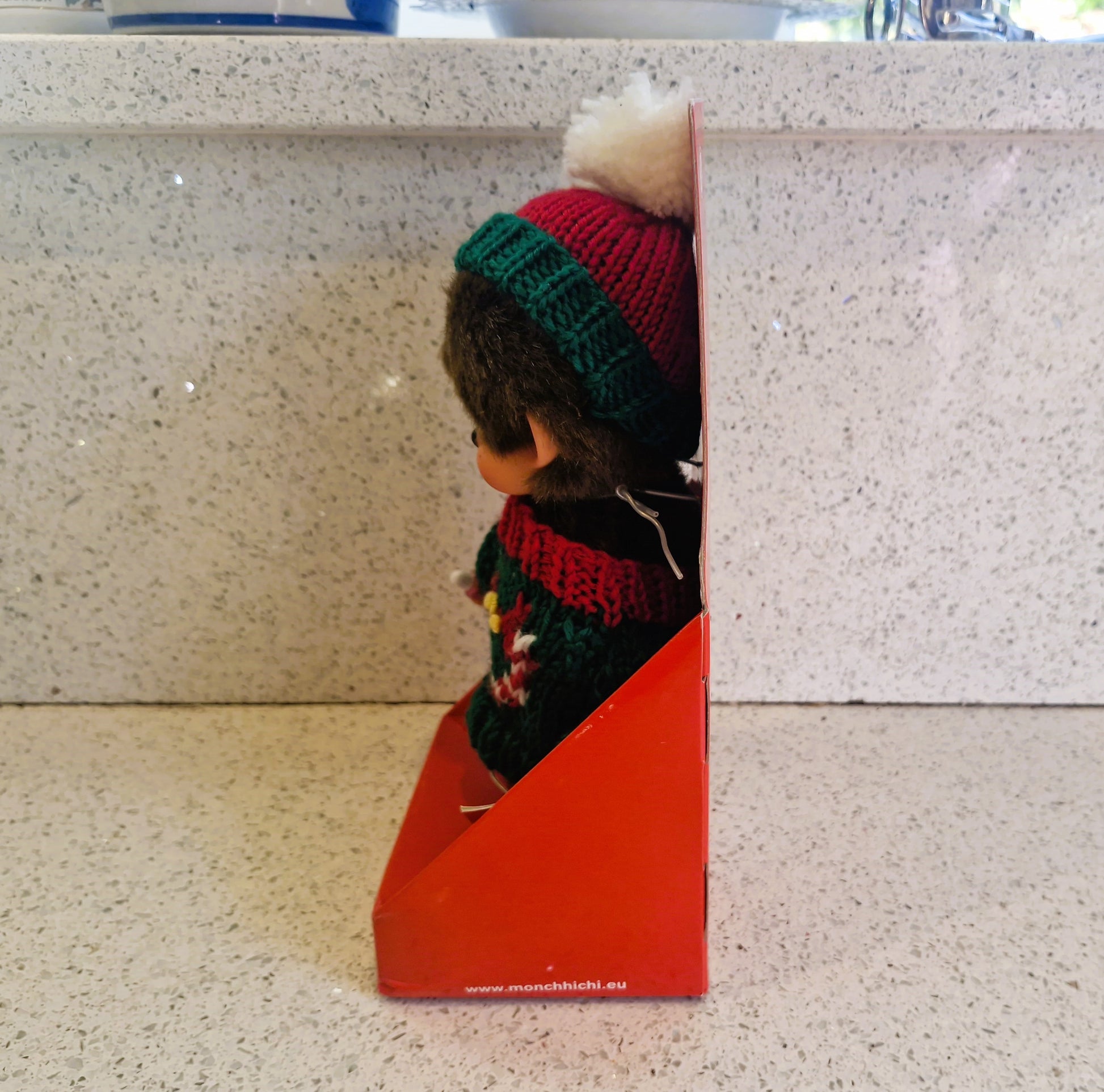Doll with colorful knitted hat and scarf peeking out from a red box on a light-colored surface.